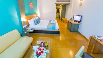 A hotel room with a bed, a desk with a computer, a sofa, and a coffee table with flowers and fruits.