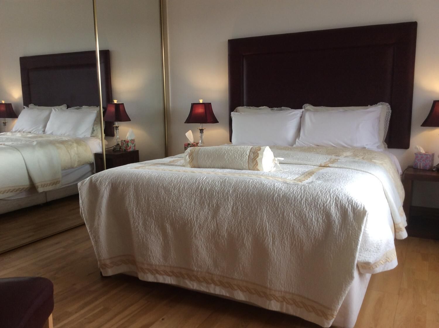 A neatly made bed with a large mirror above it, two bedside lamps, and a wooden floor.