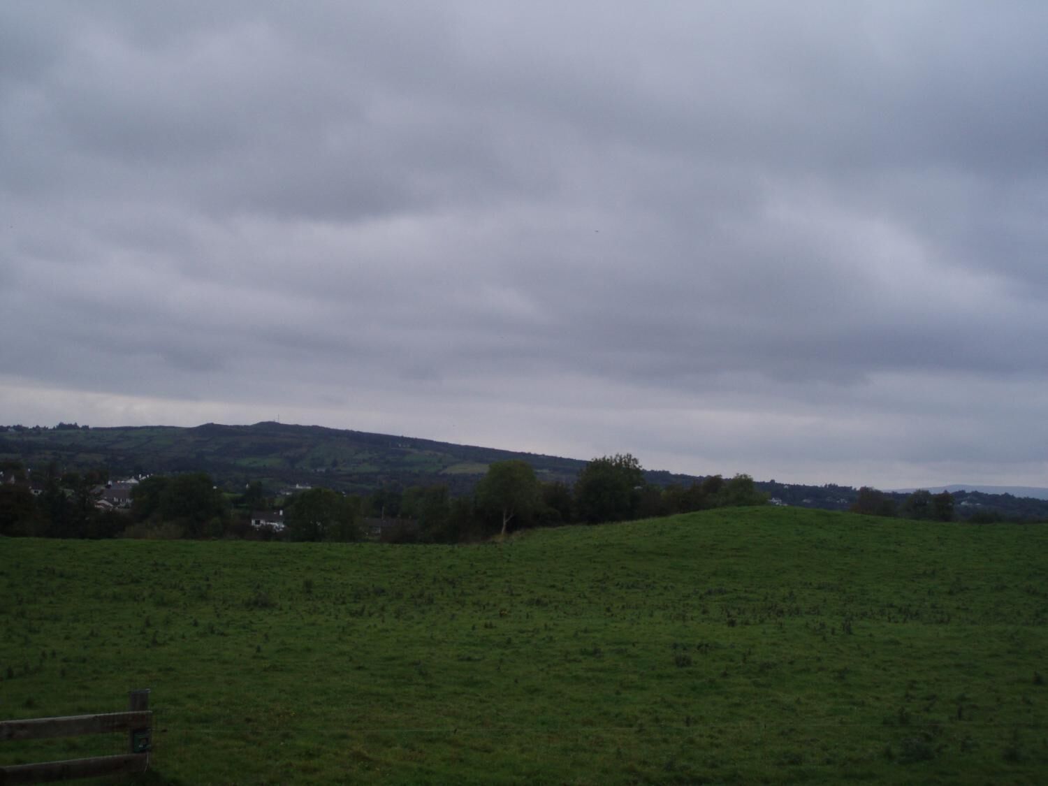 A grassy field with a wooden fence in the foreground and a hilly landscape in the background.