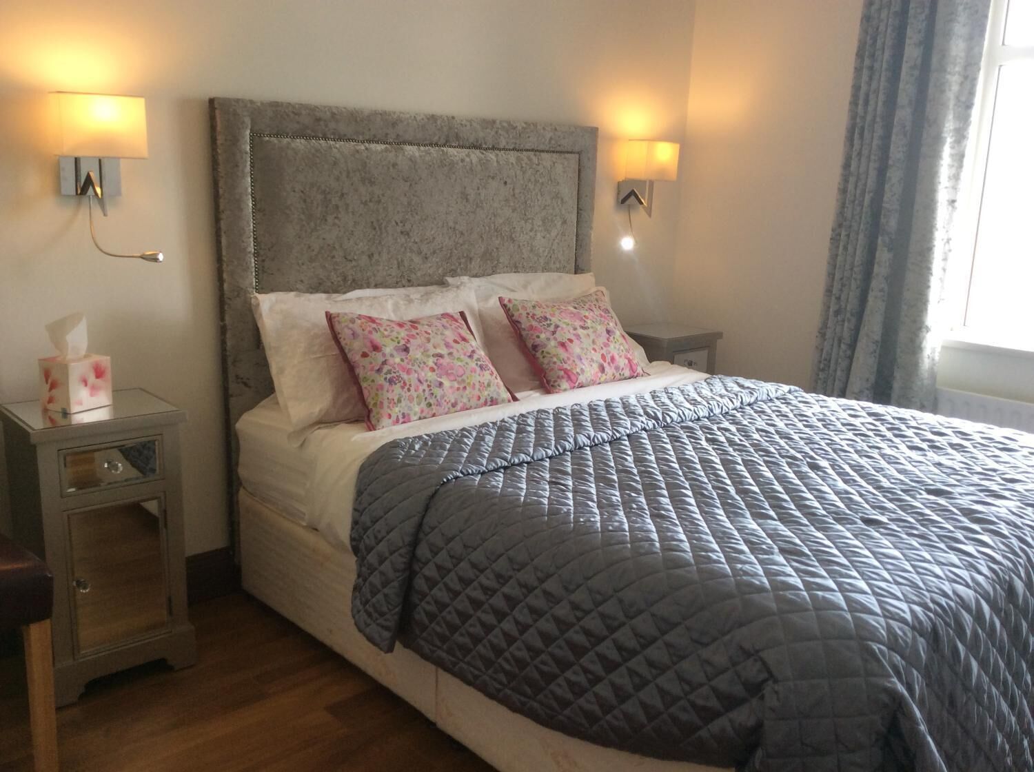 Una cama bien hecha con colcha gris, dos almohadas florales y cabecera gris. Hay lámparas de noche y una mesita de noche con una caja de pañuelos.