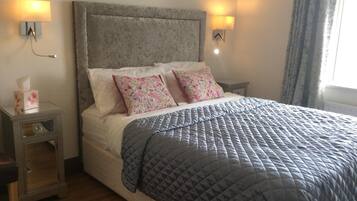 Una cama bien hecha con colcha gris, dos almohadas florales y cabecera gris. Hay lámparas de noche y una mesita de noche con una caja de pañuelos.