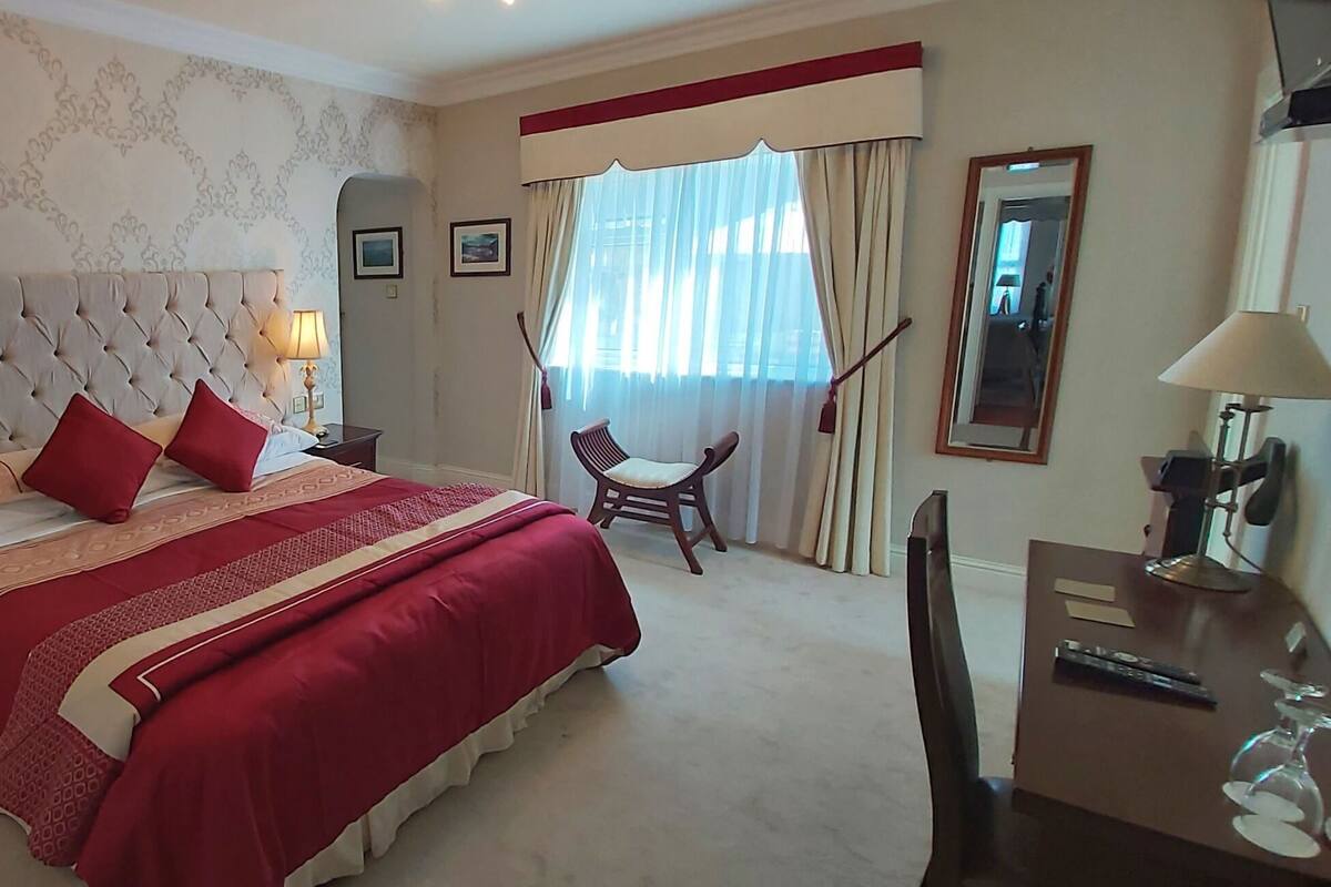 A hotel room with a large bed, two bedside lamps, a wooden nightstand, a desk with a chair, and a large window with curtains.