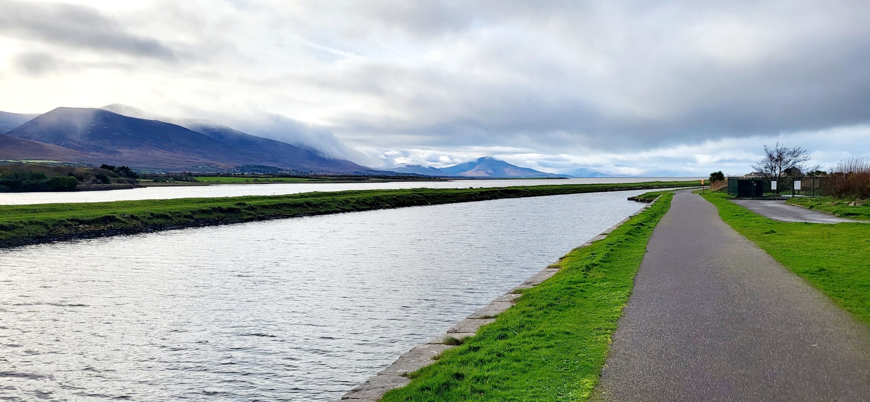 A river with a paved walkway alongside it, a grassy area, and mountains in the background.