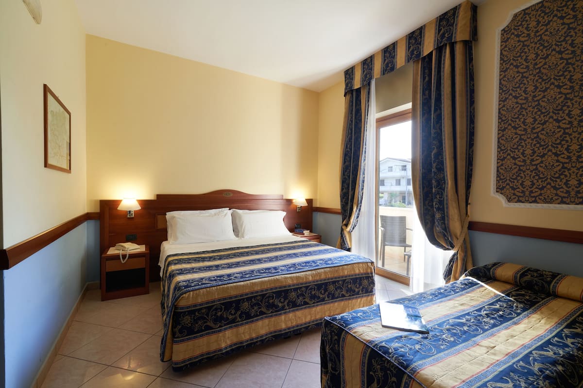 Une chambre d’hôtel avec un grand lit, un lit plus petit, une table de chevet, une commode et une fenêtre avec des rideaux.