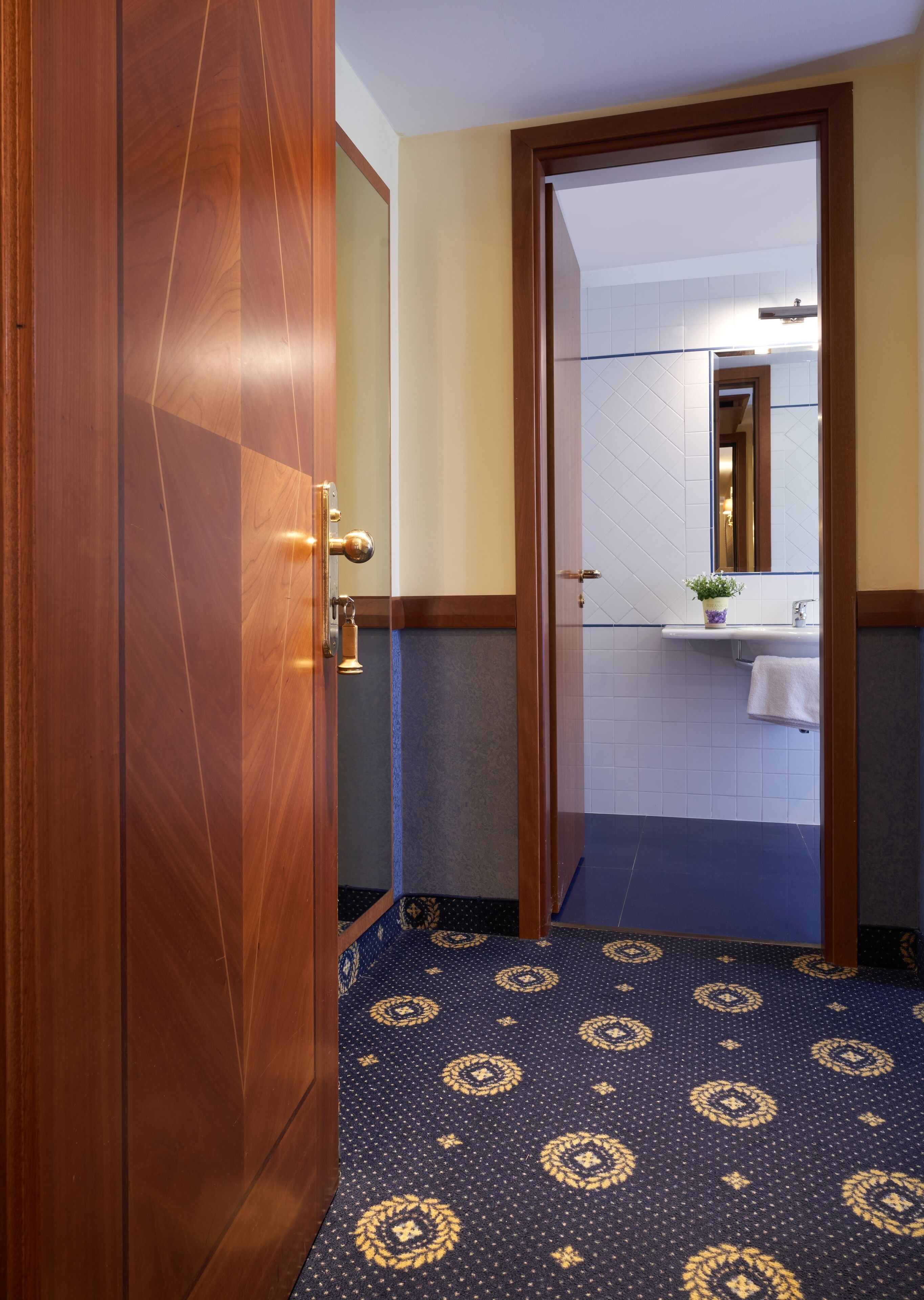 A hotel bathroom with a wooden door, a sink with a mirror, and a patterned floor.