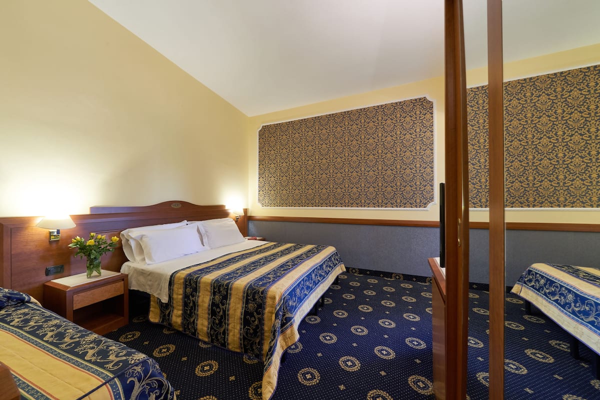 Une chambre d’hôtel avec un lit, une table de chevet avec une lampe, une fenêtre avec des rideaux à motifs et un tapis à motifs géométriques.