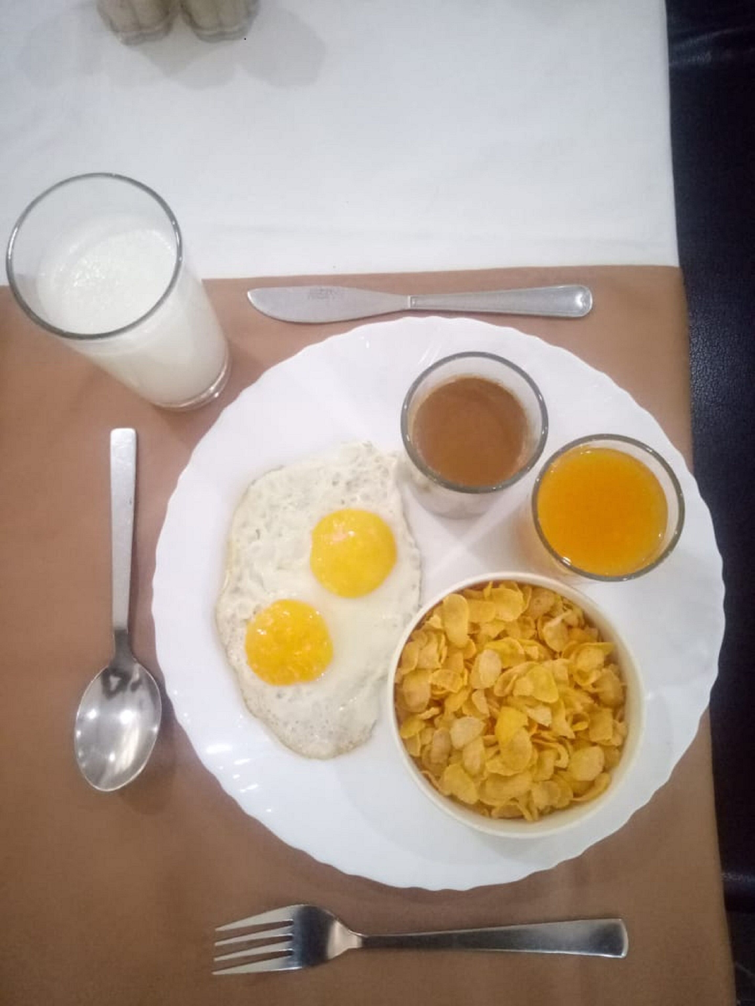 A breakfast plate with two fried eggs, a glass of milk, a small bowl of cereal, and a cup of coffee.