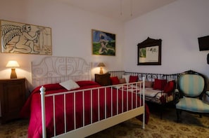 Un dormitorio con estructura de cama de metal blanco, ropa de cama roja, una mesita de noche de madera con lámpara, una silla y decoración en la pared.