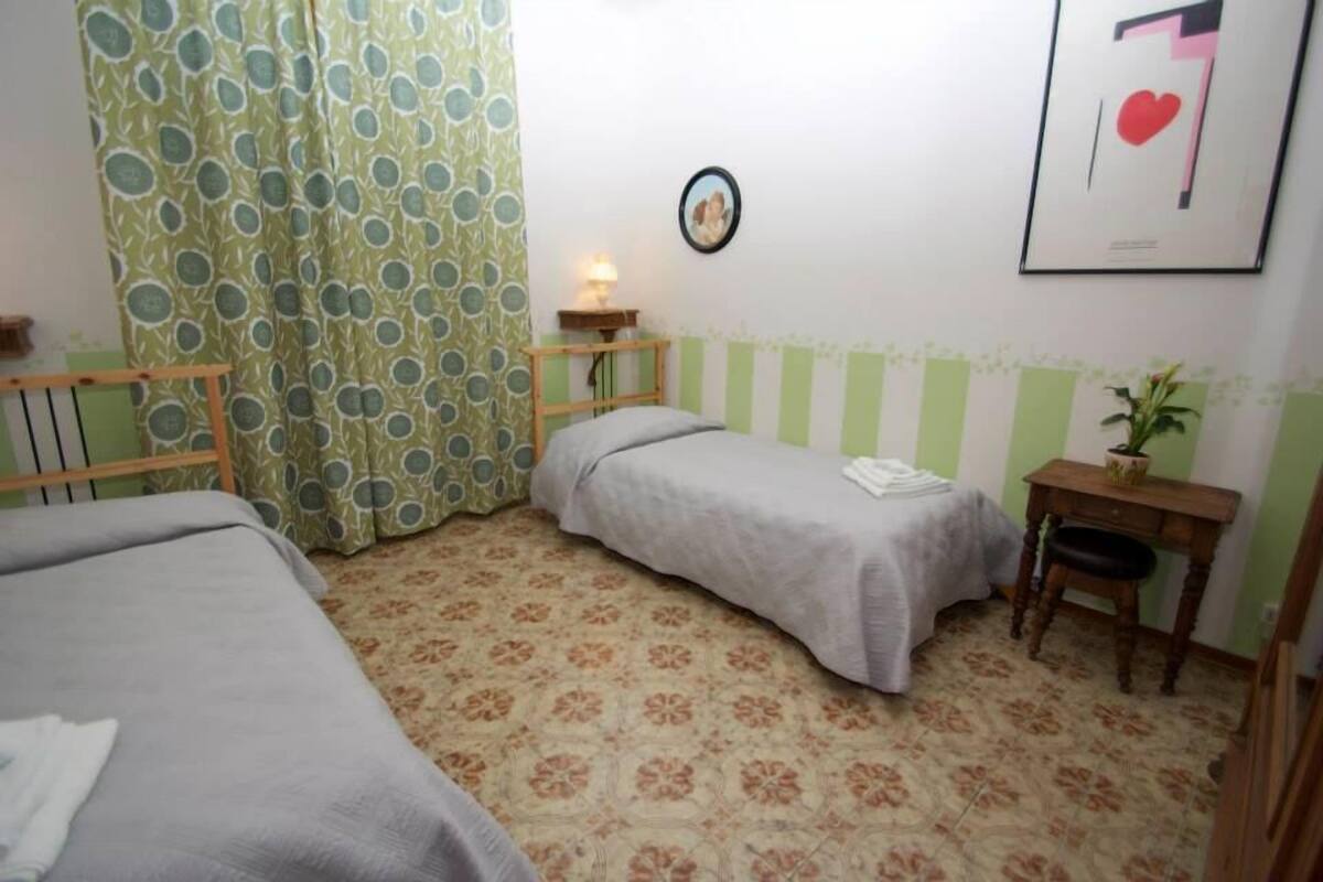 Une petite chambre agréable et confortable, avec deux lits simples, un bureau, une chaise, une petite table, une lampe et un tableau encadré au mur.