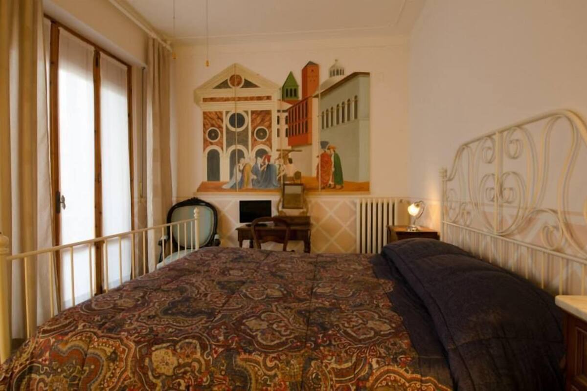 Une chambre à coucher avec une couvre-lit à motifs, une tête de lit en métal, un bureau avec une chaise et un tableau mural.