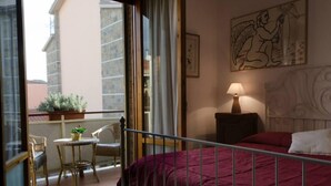A hotel room with a balcony, a bed with a red bedspread, a small table with a plant, a chair, and a nightstand with a lamp. - Bed and Breakfast La Casa Di Elide (Arezzo)