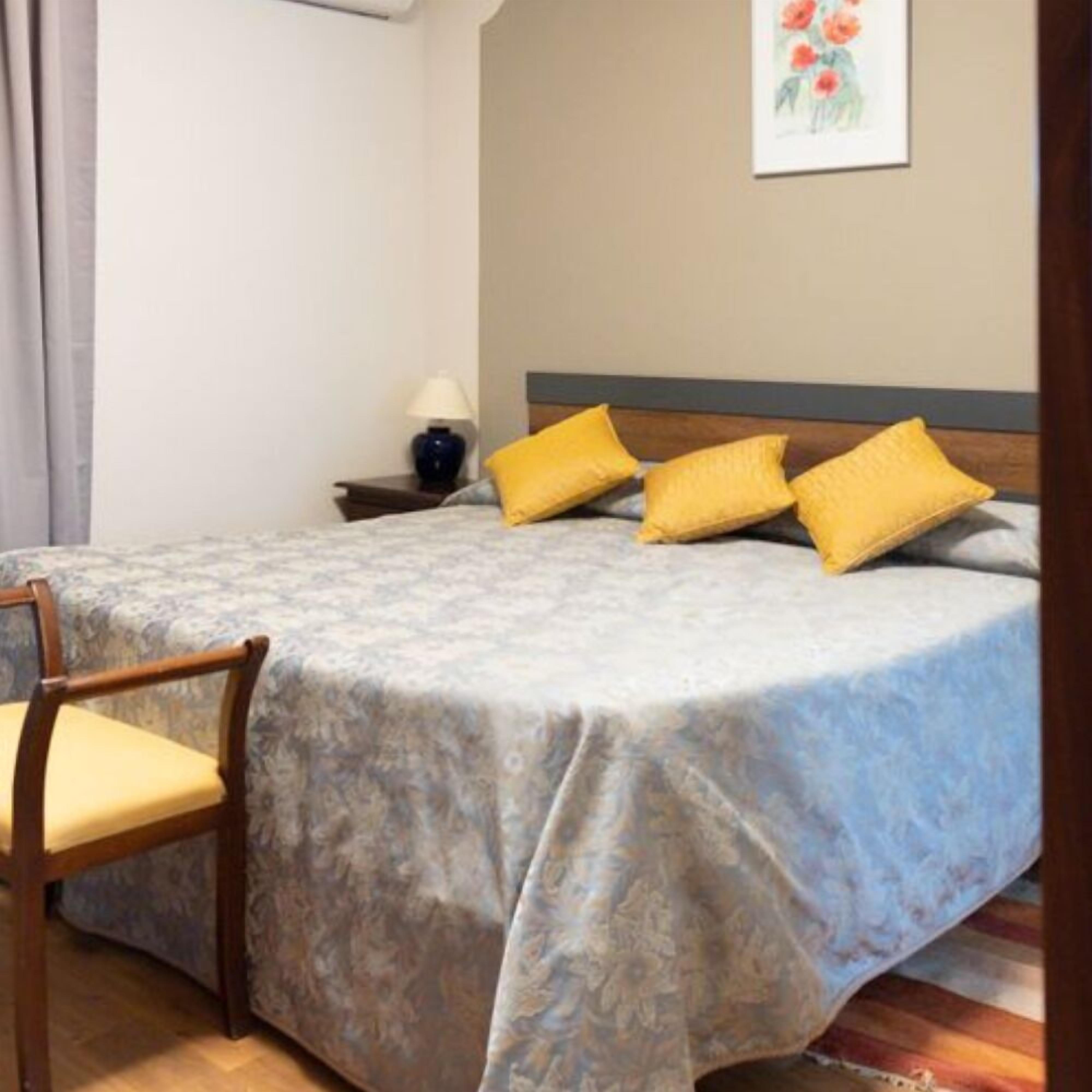 Een net opgemaakt bed met een bloemenbedekker, een houten stoel met kussen, en een nachtkastje met lamp.