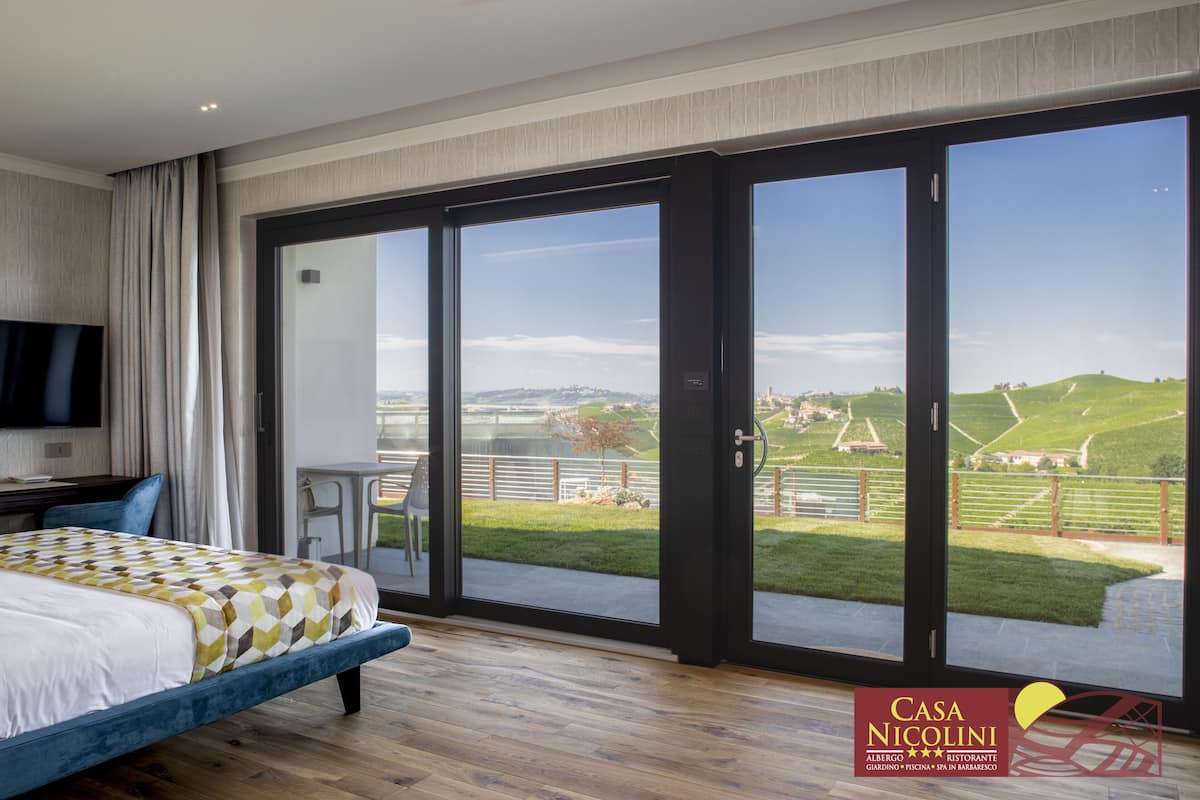 A bedroom with a large bed, a TV, and a sliding glass door leading to a balcony with a view of a green landscape.