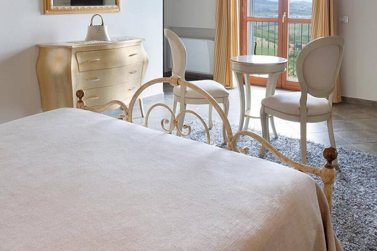 A bedroom with a bed, a dresser, a chair, a table, a mirror, and a balcony with curtains.