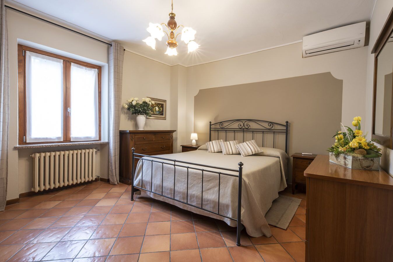 Une chambre à coucher avec un lit en métal, une commode en bois, un radiateur, une fenêtre avec des rideaux, un lustre et un climatiseur fixé au mur.