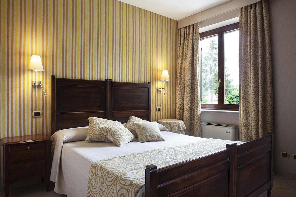 A hotel room with a large bed, two bedside tables, a nightstand, a window with curtains, and a wall-mounted heater.