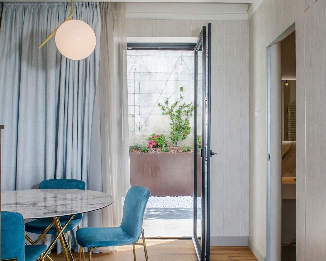 A modern dining area with a round table and blue chairs, a large window with curtains, and a view of a garden outside.