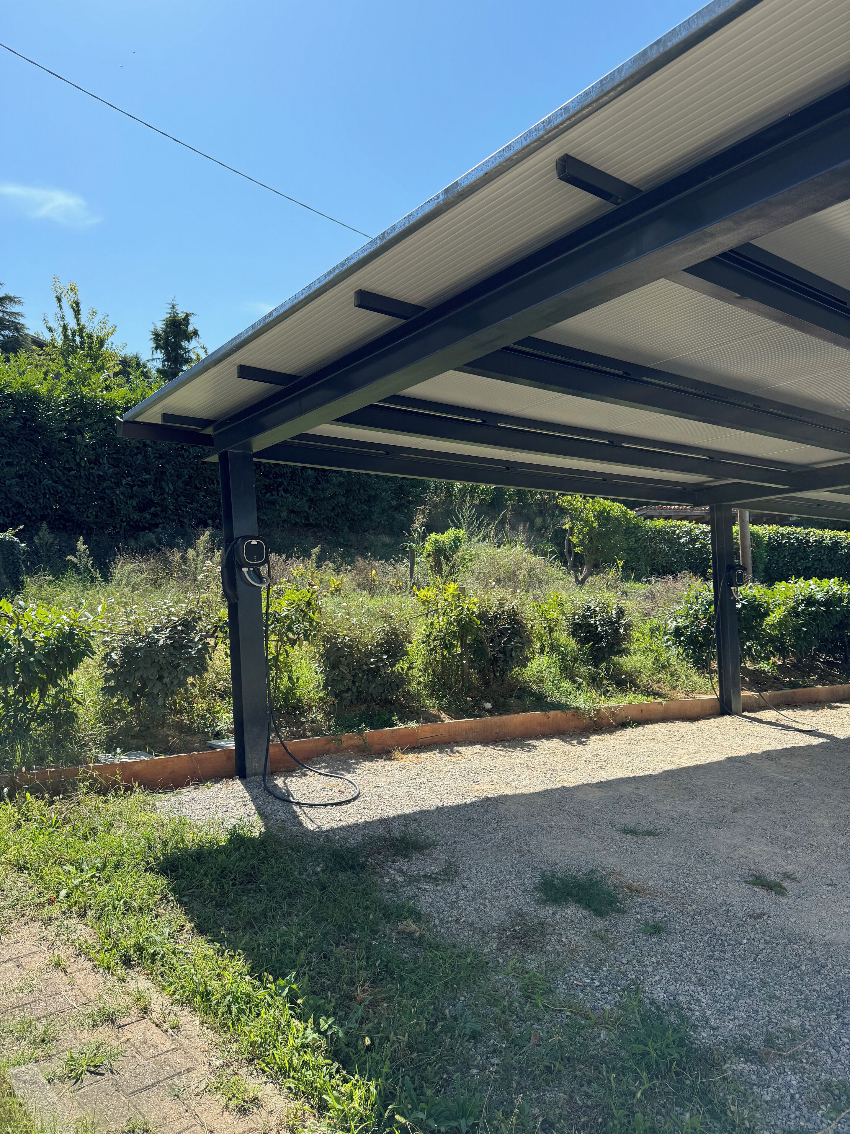 Un espacio al aire libre cubierto con un sendero de grava, rodeado de vegetación y una estructura de techo metálico.
