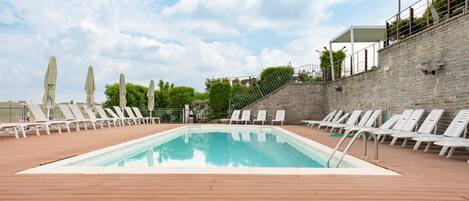 A rectangular pool with white lounge chairs and umbrellas on a wooden deck, surrounded by stone walls and greenery.