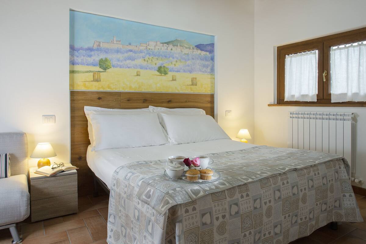 A neatly made bed with a patterned bedspread, a bedside table with a lamp, a chair, and a painting of a landscape on the wall.