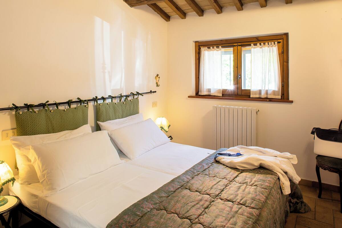 A neatly made bed with white linens, a quilted blanket, and a wooden headboard. There is a window with curtains, a radiator, and a chair to the right.