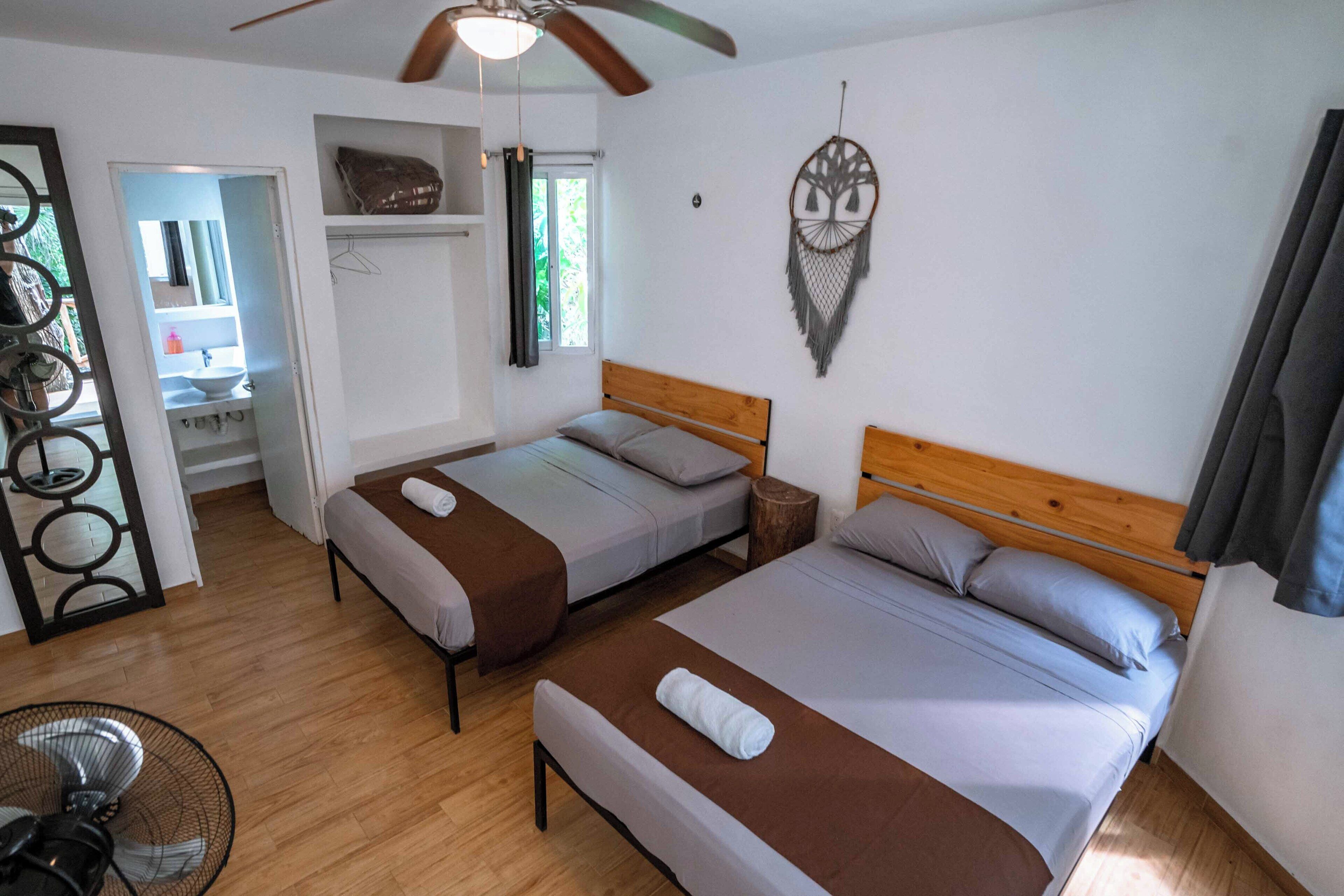 A room with two beds, a ceiling fan, a bedside table, a wall-mounted dreamcatcher, and a window with curtains.