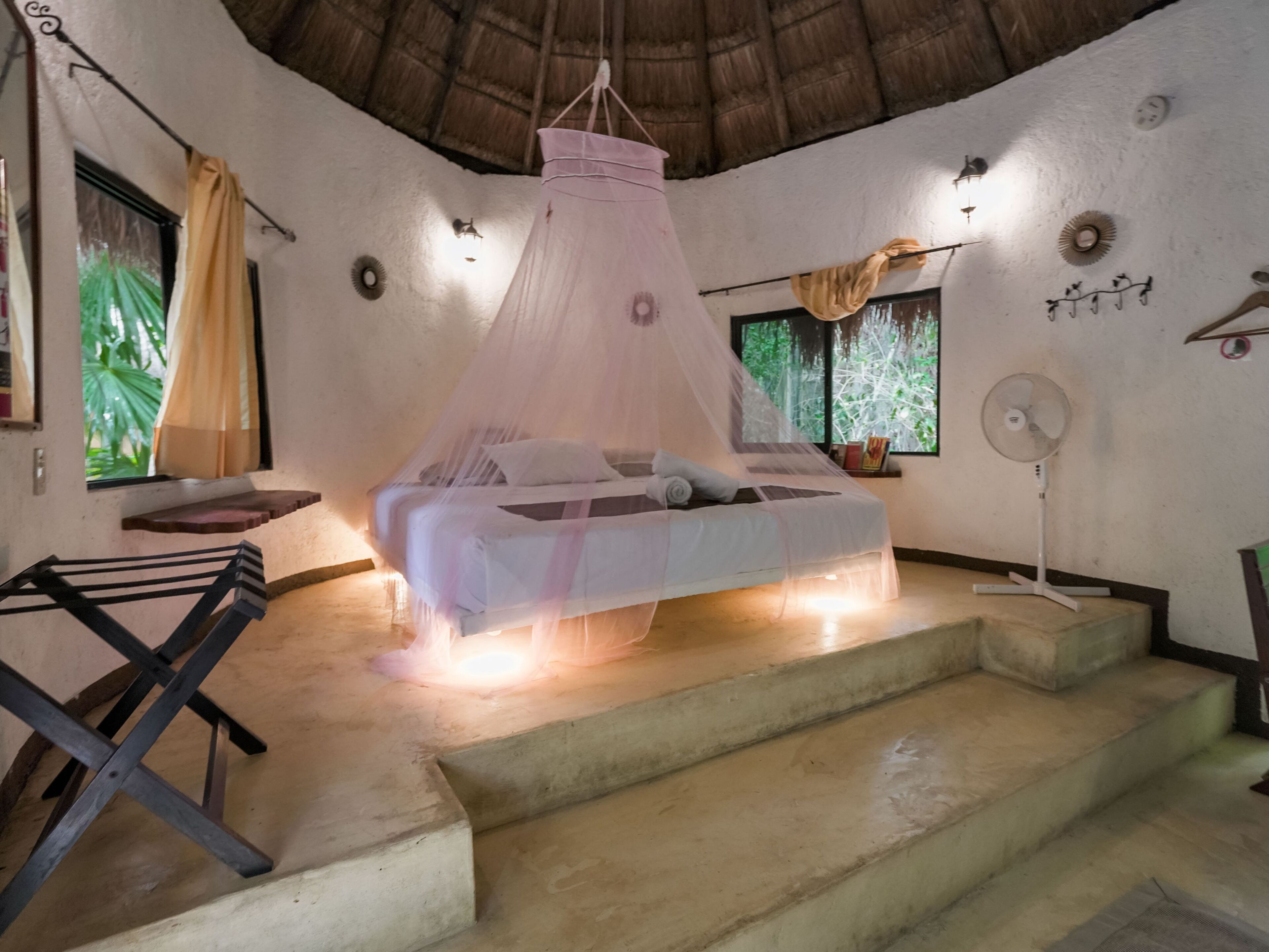 Un dormitorio con una cama bajo una mosquitera, un ventilador, una ventana con cortinas y una escalera.