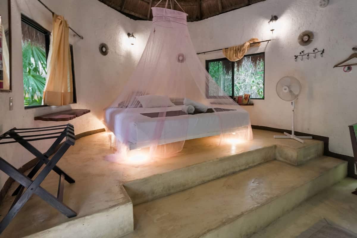 Une chambre à coucher avec un lit recouvert d’une moustiquaire, un ventilateur, une fenêtre avec des rideaux et un escalier.