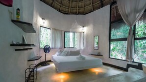 A bedroom with a bed, bedside tables, a standing fan, and large windows with curtains. - Jolie Jungle Eco Hotel (Leona Vicario)