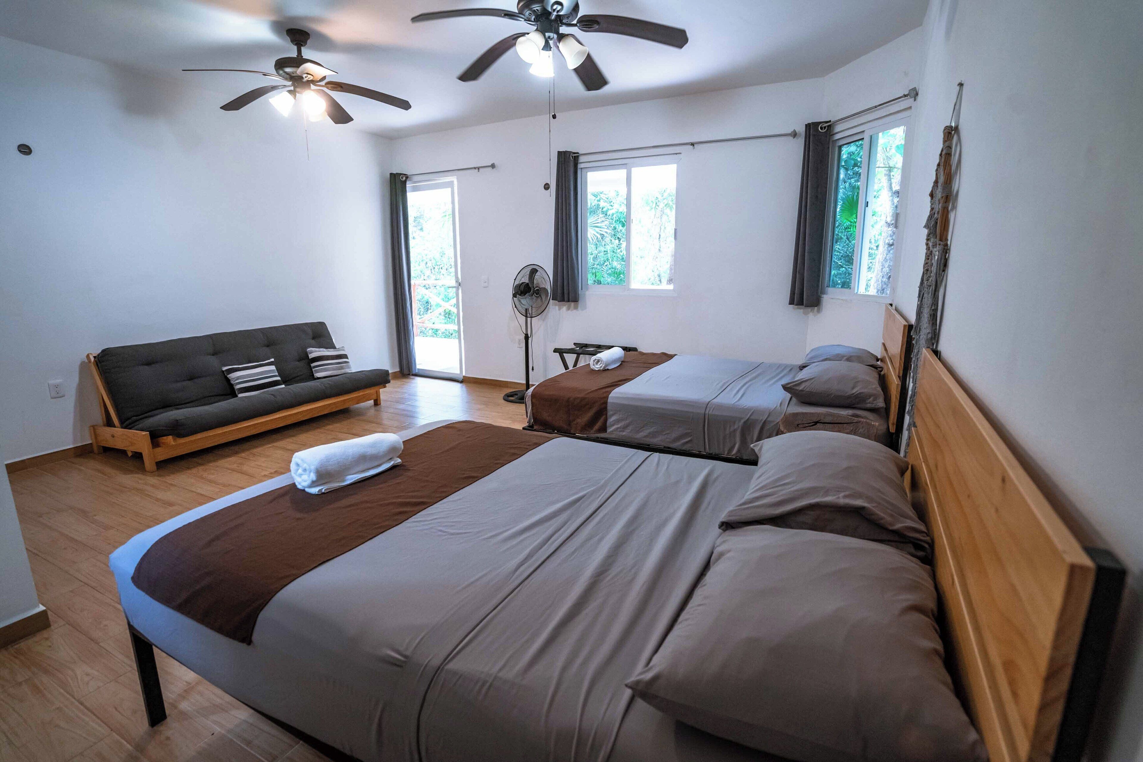 A bedroom with two beds, a sofa, ceiling fans, and large windows.