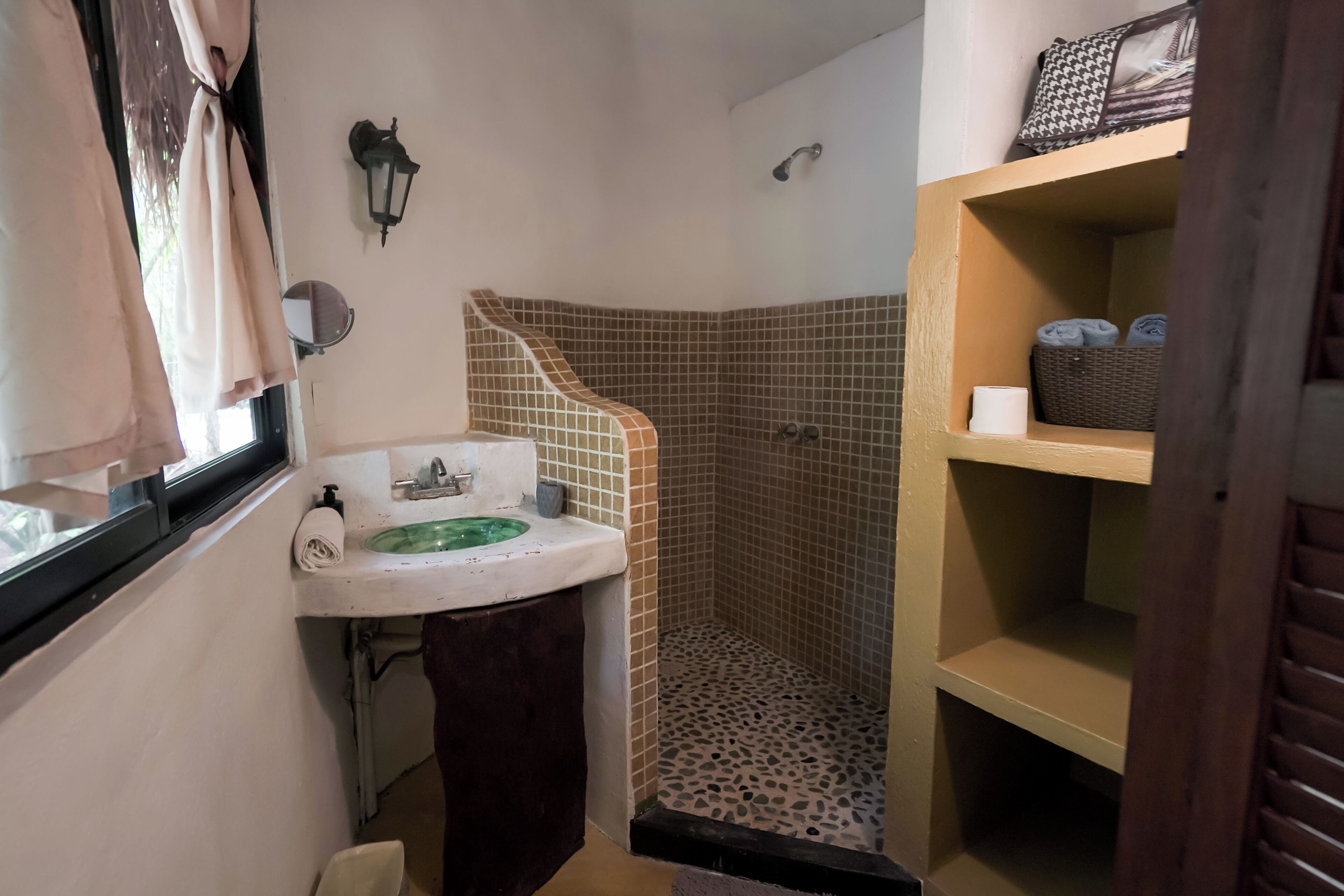 Casa de banho com lavatório, espelho, chuveiro e chão em azulejo, e uma prateleira com toalhas.