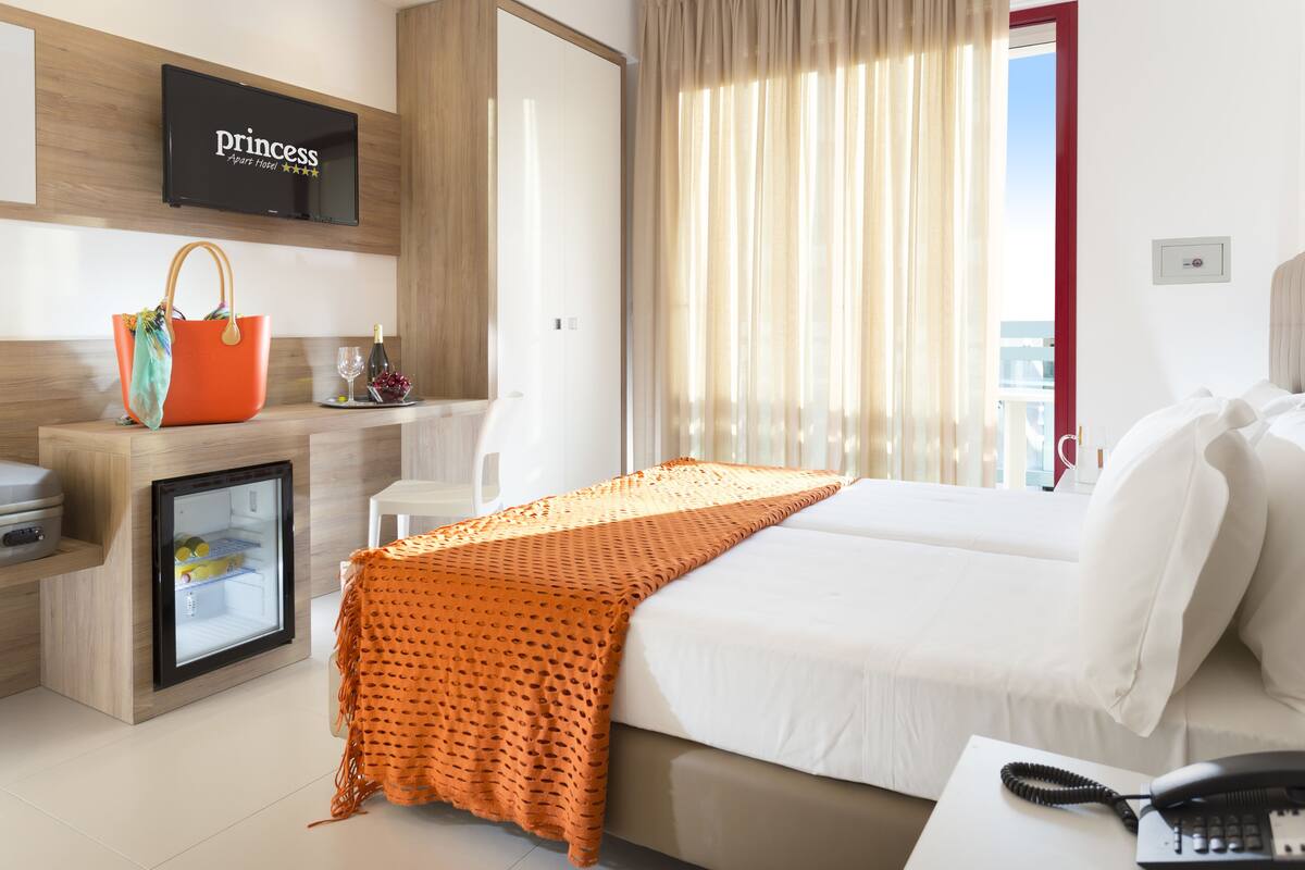 Camera d'albergo con un letto, un comodino con il telefono, un mini-frigorifero, un minibar, una TV a parete e una finestra con tende.