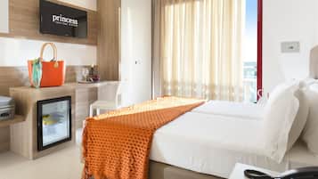 Ein Hotelzimmer mit einem Bett, einem Nachttisch mit Telefon, einem Kühlschrank, einer Minibar, einem an der Wand befestigten Fernseher und einem Fenster mit Vorhängen.