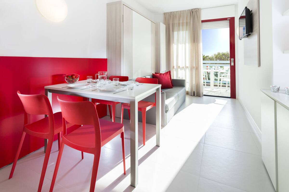 Un'area pranzo moderna con un tavolo bianco e sedie rosse, un balcone con ringhiera rossa e vista sul verde.