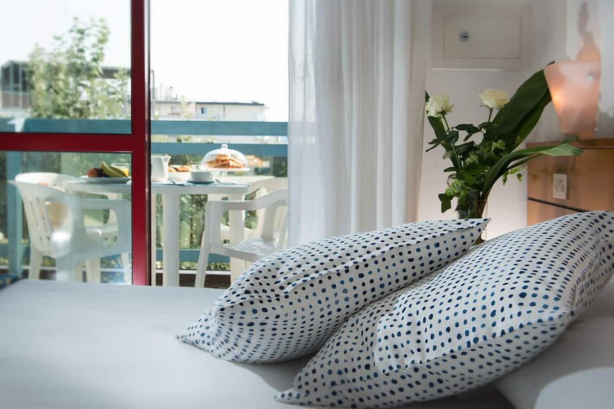 Una camera d'hotel con un letto, cuscini bianchi a pois, un vaso con fiori bianchi e un balcone con un tavolo apparecchiato per la colazione.