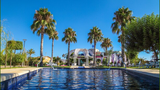 A swimming pool with a clear blue water surface, surrounded by palm trees and a building with a dome-like structure in the background. - Grand Hotel Paclà (Avola)