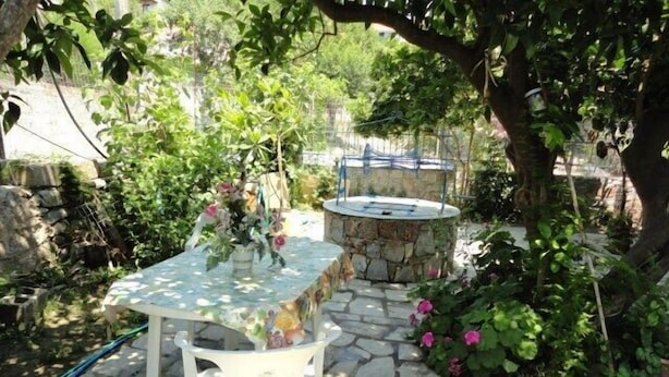 Un patio de piedra con una mesa y una silla blancas, rodeado de follaje y un muro de piedra.