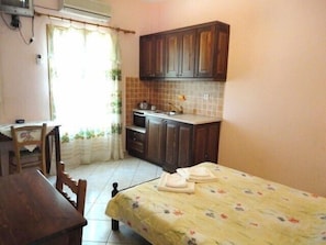A small, compact room with a bed, a desk, a chair, a kitchenette, and a window with curtains.