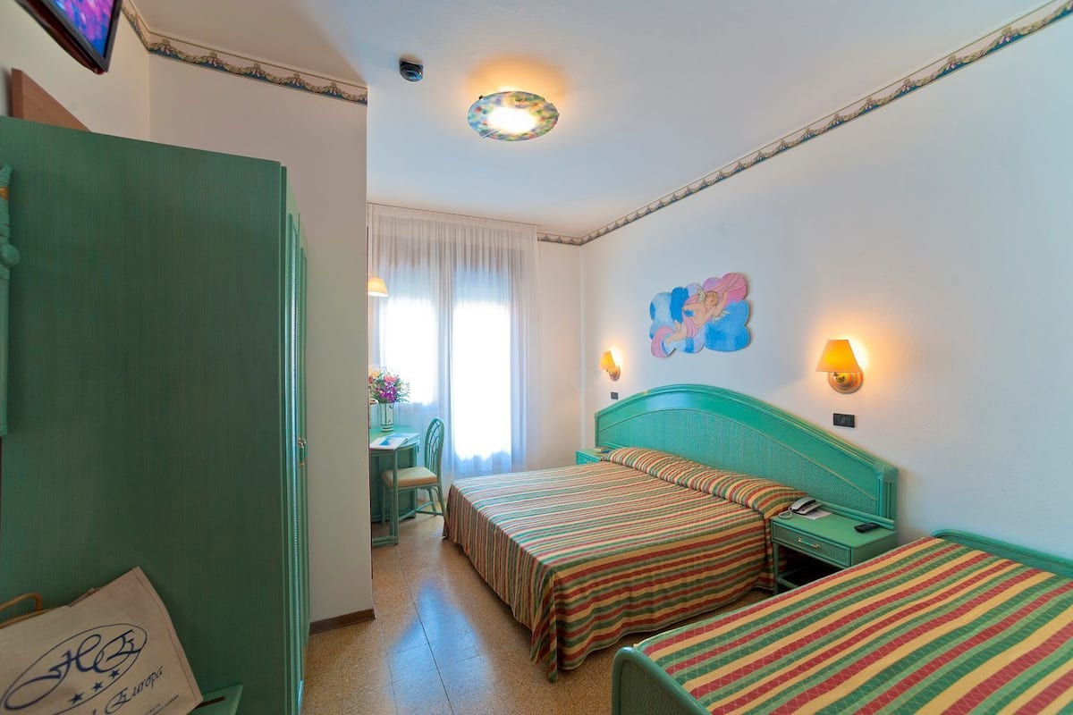 Une chambre d’hôtel avec deux lits simples, un bureau avec une chaise, une fenêtre avec des rideaux et un tableau décoratif au mur.