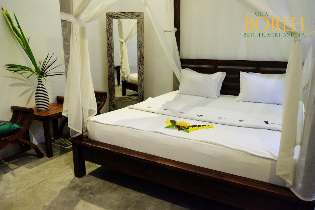 A neatly made bed with white linens and a decorative flower, a wooden nightstand with a striped vase, and a mirror with a decorative frame.
