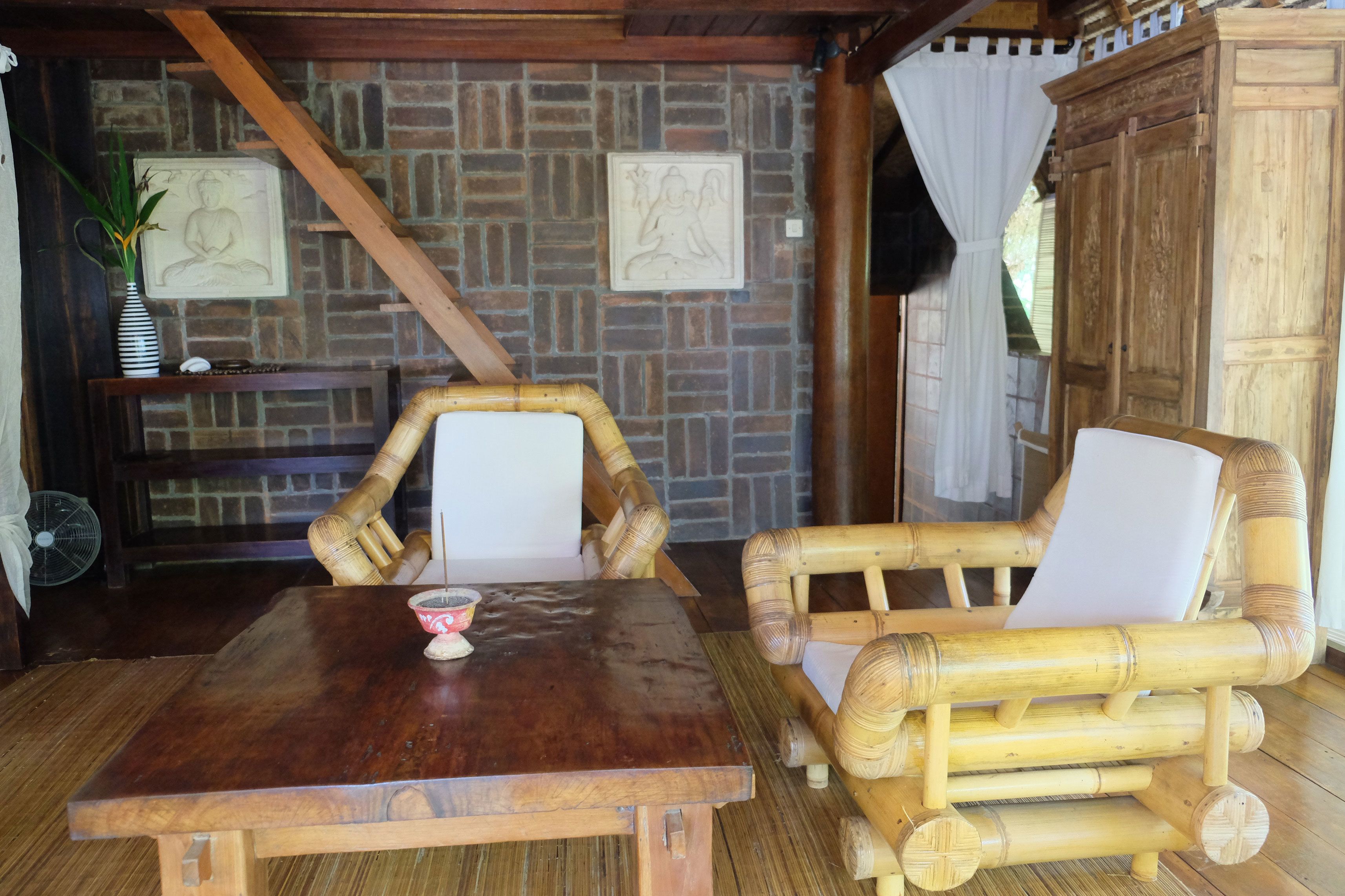 Une table en bois, deux chaises en bambou, un petit objet décoratif et un mur en briques orné de deux tableaux encadrés.