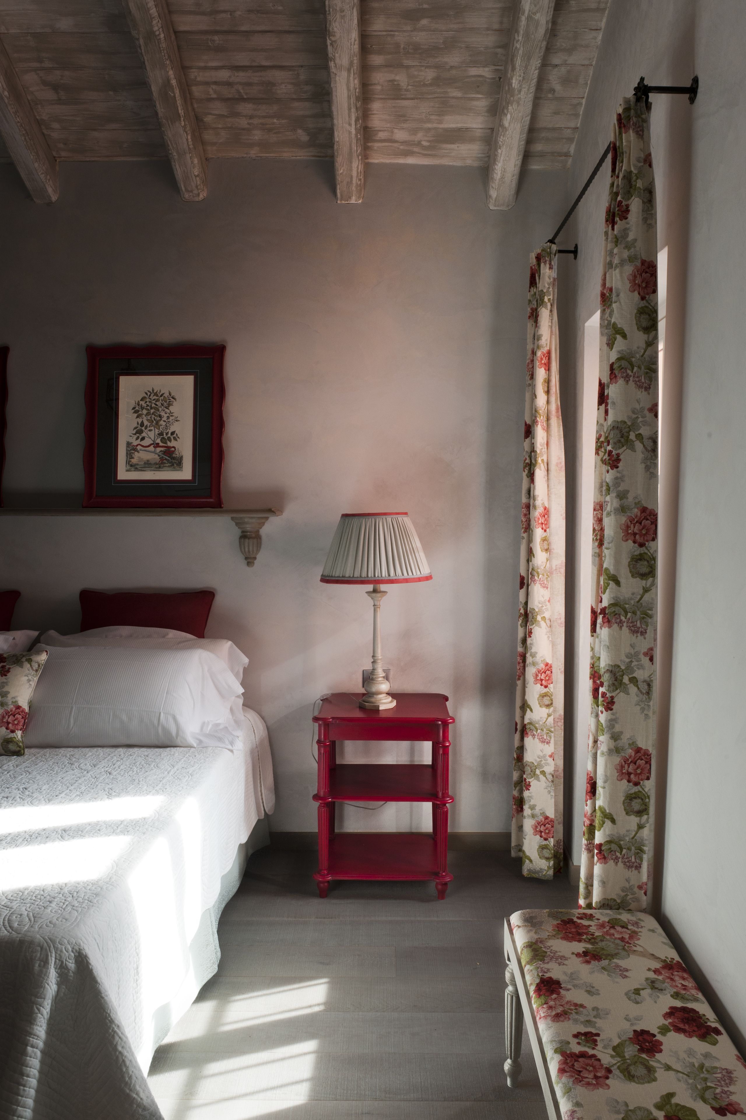A bedroom with a bed, a nightstand with a lamp, a framed picture on the wall, and floral curtains.
