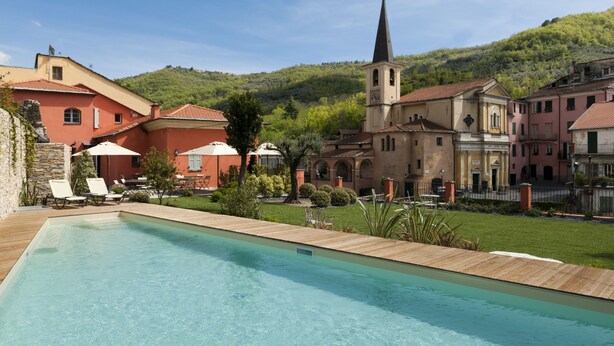 A swimming pool with lounge chairs, a church, and buildings with red and beige facades. - Relais Del Maro (Borgomaro)