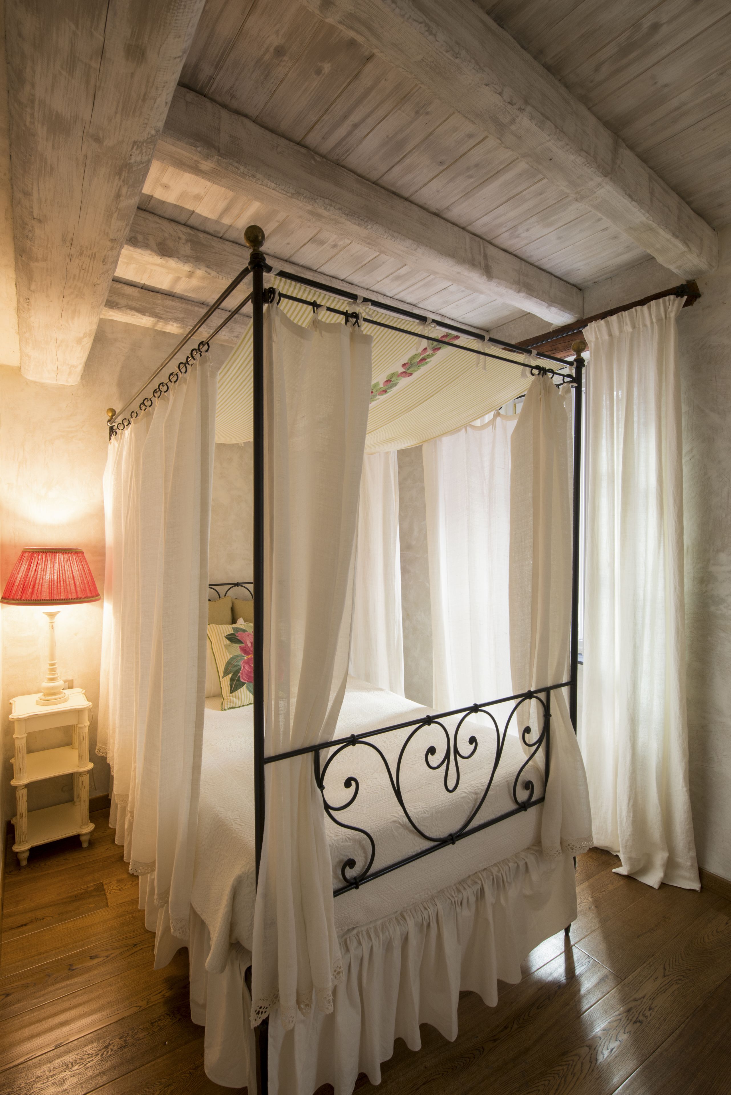 A four-poster bed with white curtains, a bedside table with a lamp, and wooden flooring.