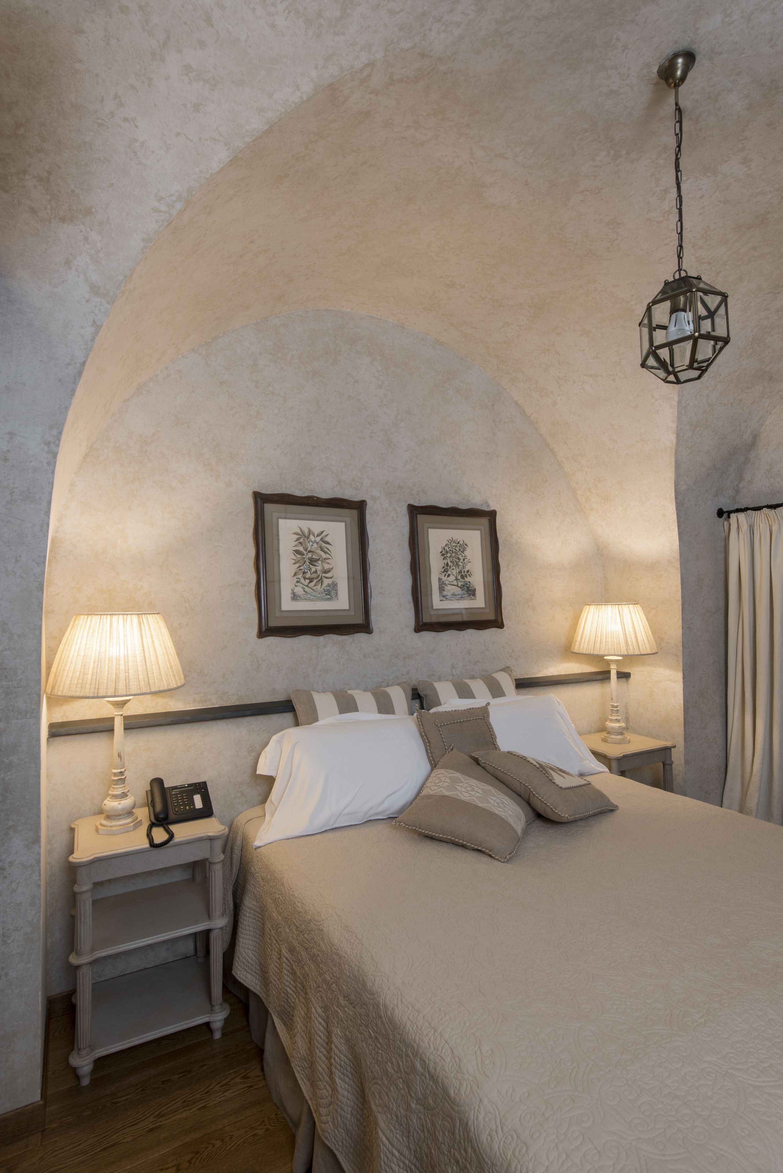 Un dormitorio con una cama grande, dos mesitas de noche con lámparas, un teléfono en una de las mesitas y cuadros enmarcados en la pared.