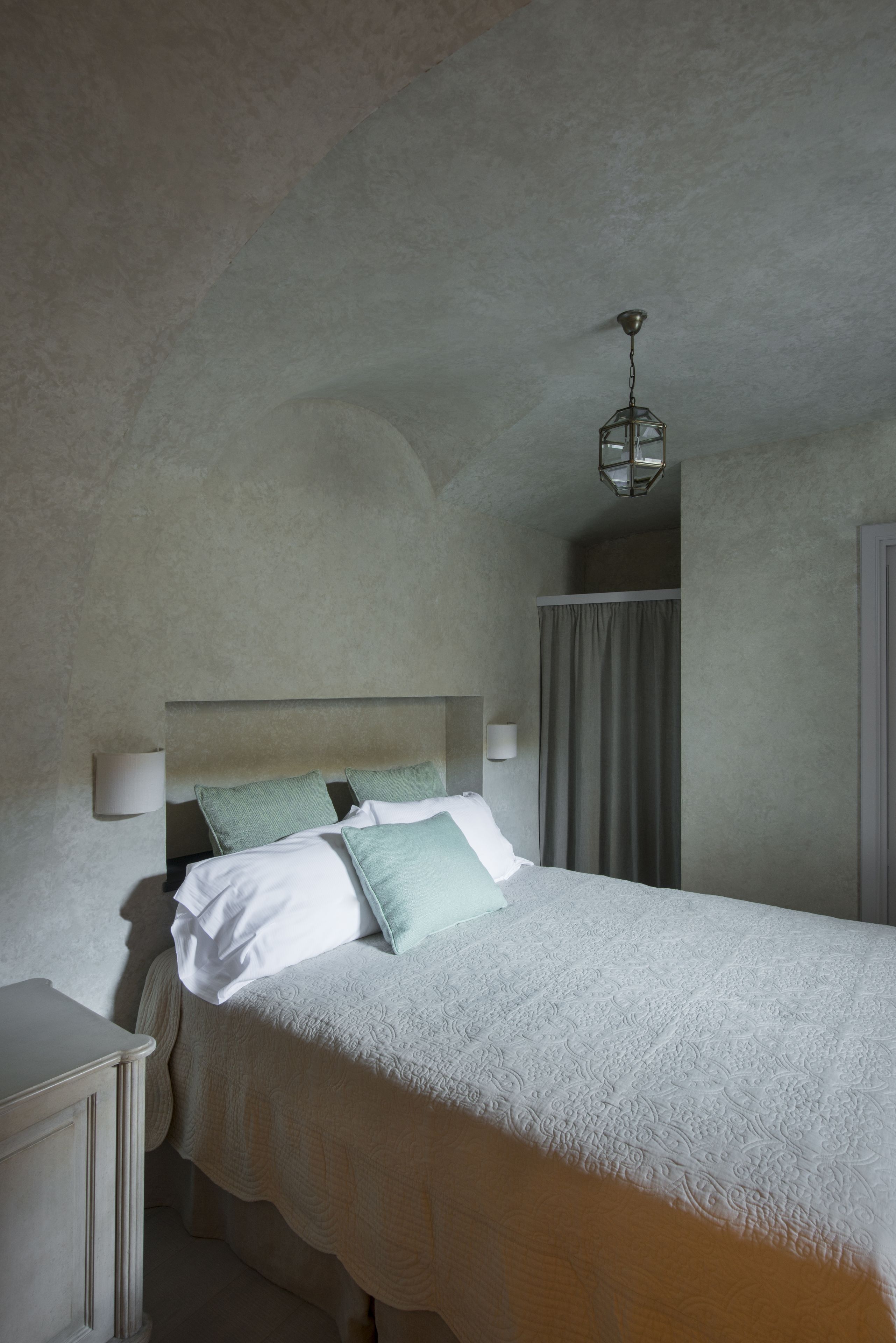 Une chambre à coucher avec un lit, une table de chevet, un luminaire suspendu et une fenêtre avec des rideaux.