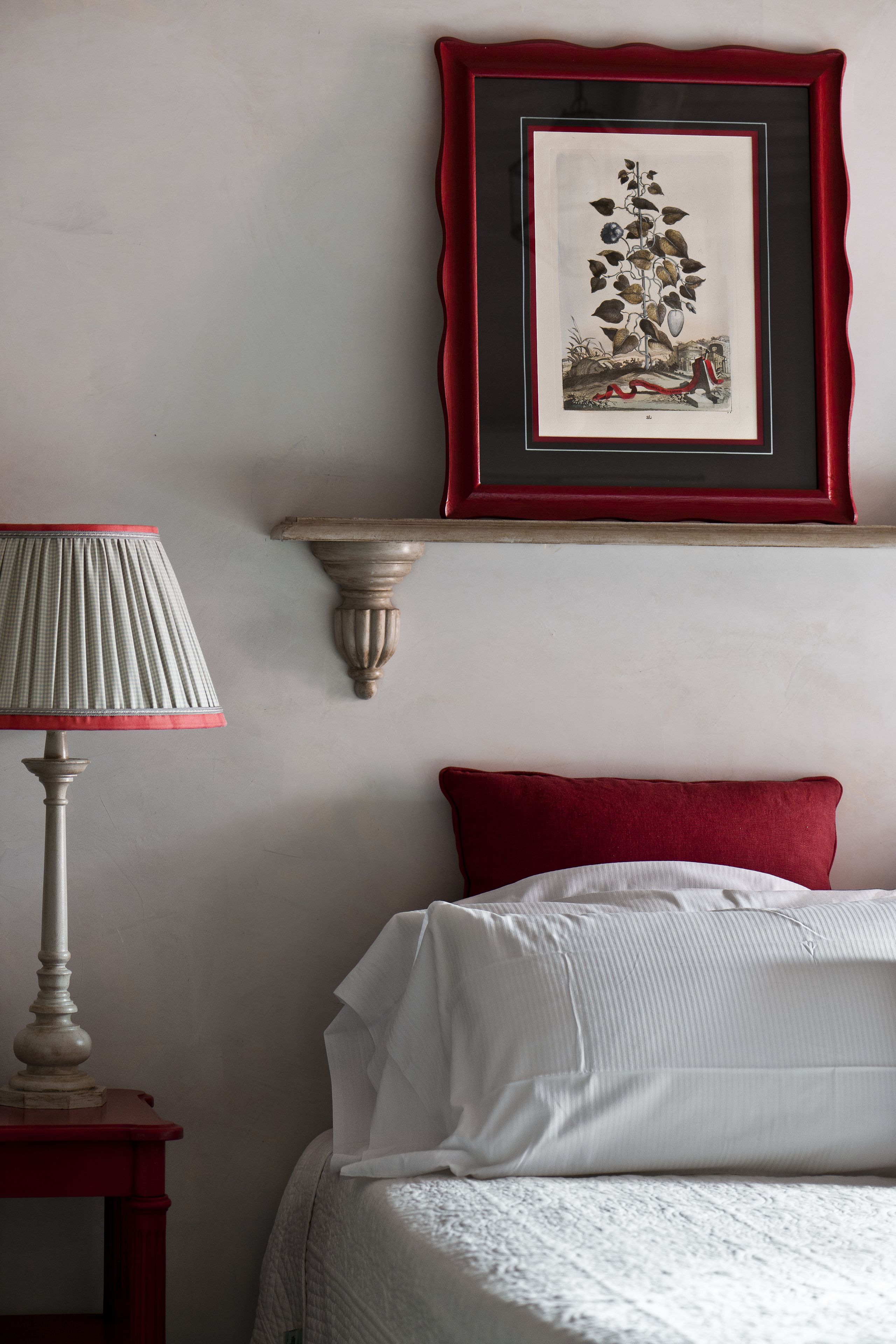 Una cama bien hecha con un almohadón rojo, una lámpara blanca sobre una mesita de noche de madera, e ilustración botánica enmarcada en la pared.