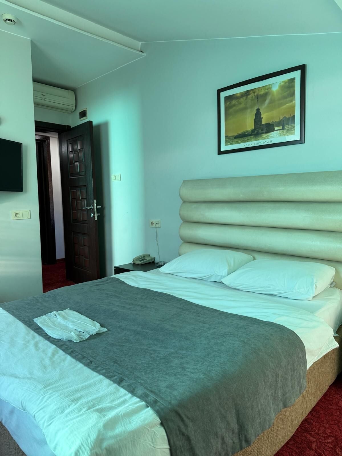A hotel room with a large bed, a bedside table with a lamp, a framed picture on the wall, and a door leading to another room.