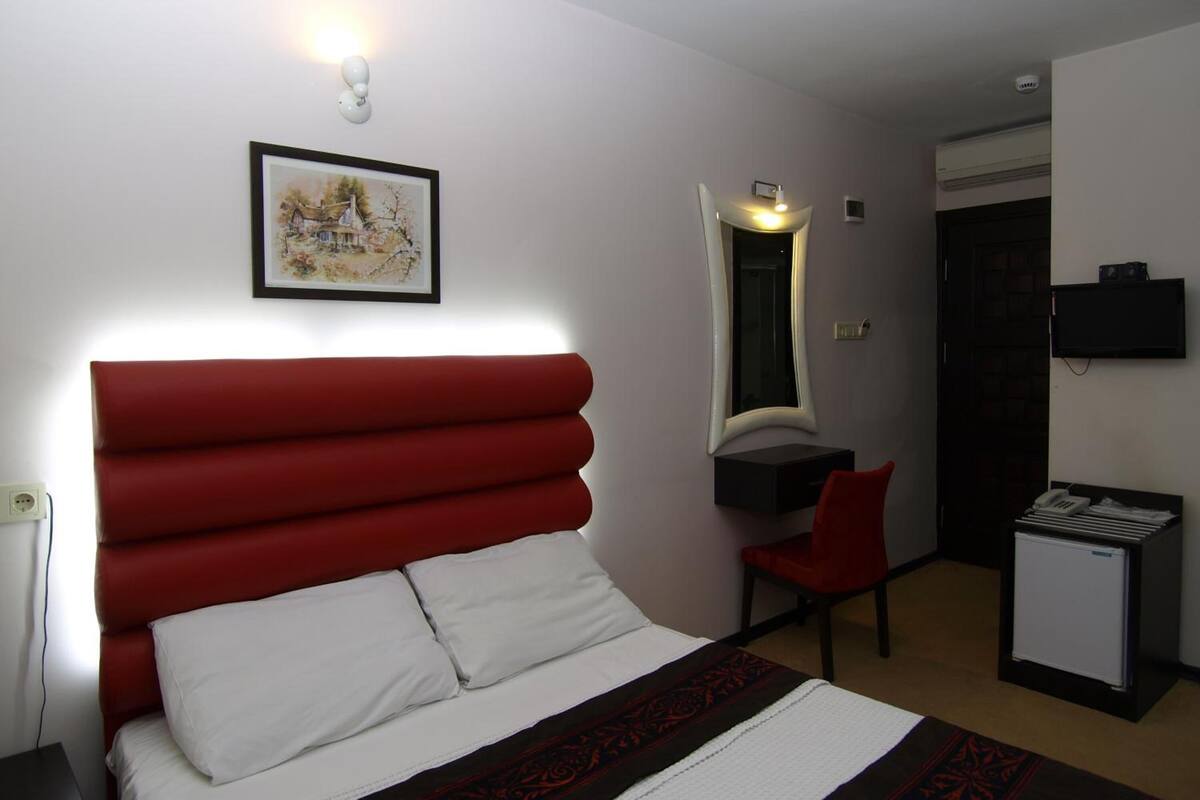 Ein Hotelzimmer mit rotem, gepolsterten Kopfteil, weißer Bettdecke, einem gerahmten Bild an der Wand, einem Schreibtisch mit Stuhl, einem kleinen Kühlschrank und einem flachbildfernseher, der an der Wand befestigt ist.