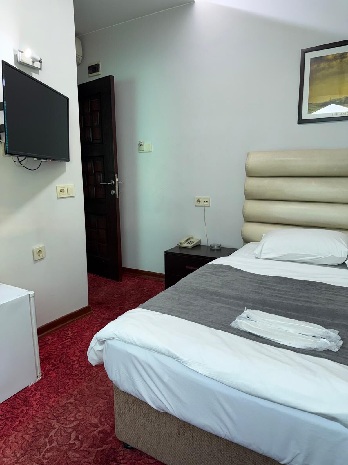 Ett hotellrum med en säng, en tv som är monterad på väggen, ett nattduksbord med en telefon och ett rött heltäckningsmatta på golvet.